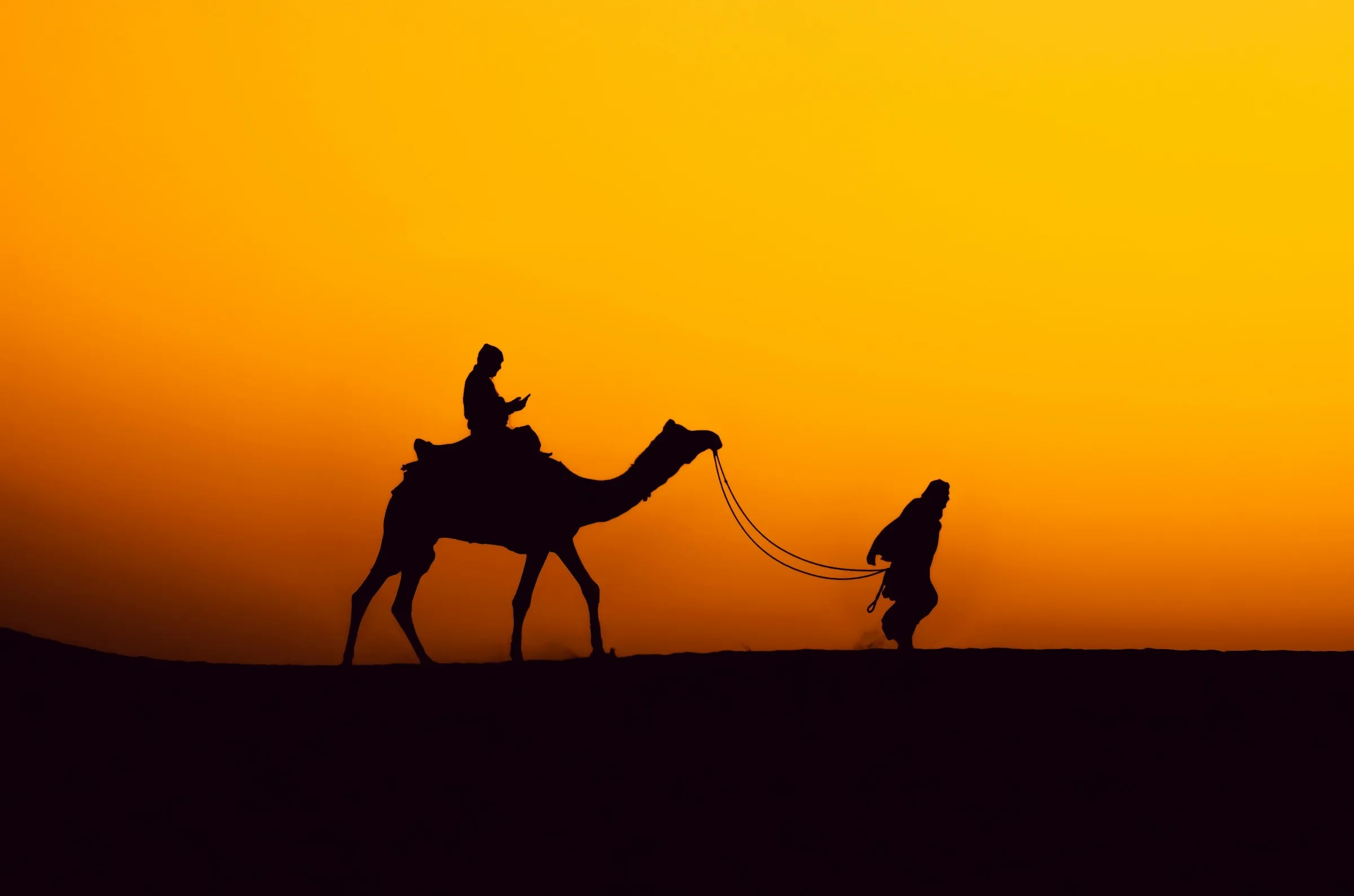 A camel and its herder silhouetted against an orange Thar Desert sunset