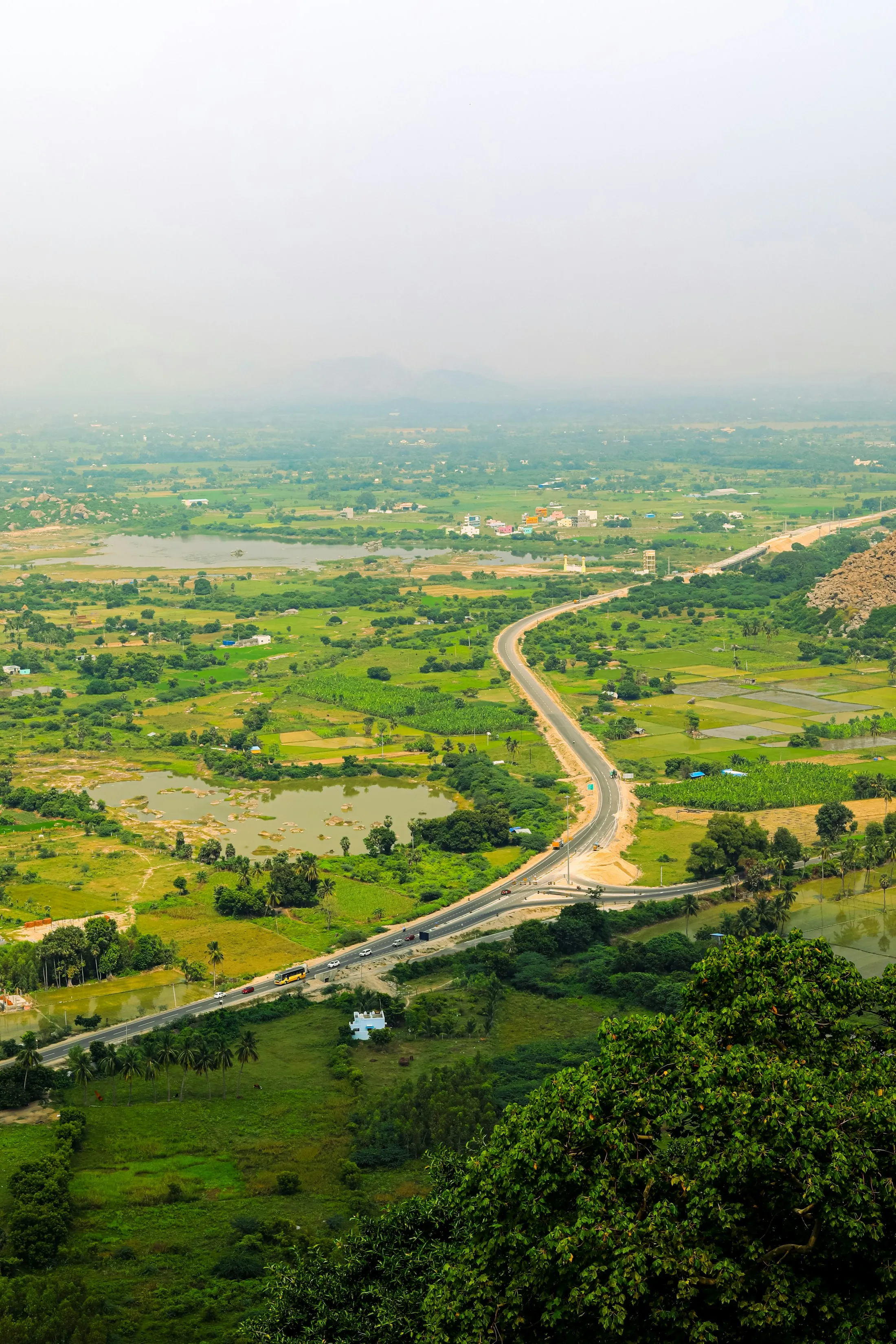 An elevated view of a highway winding through a green valley of fields towards distant hazy hills